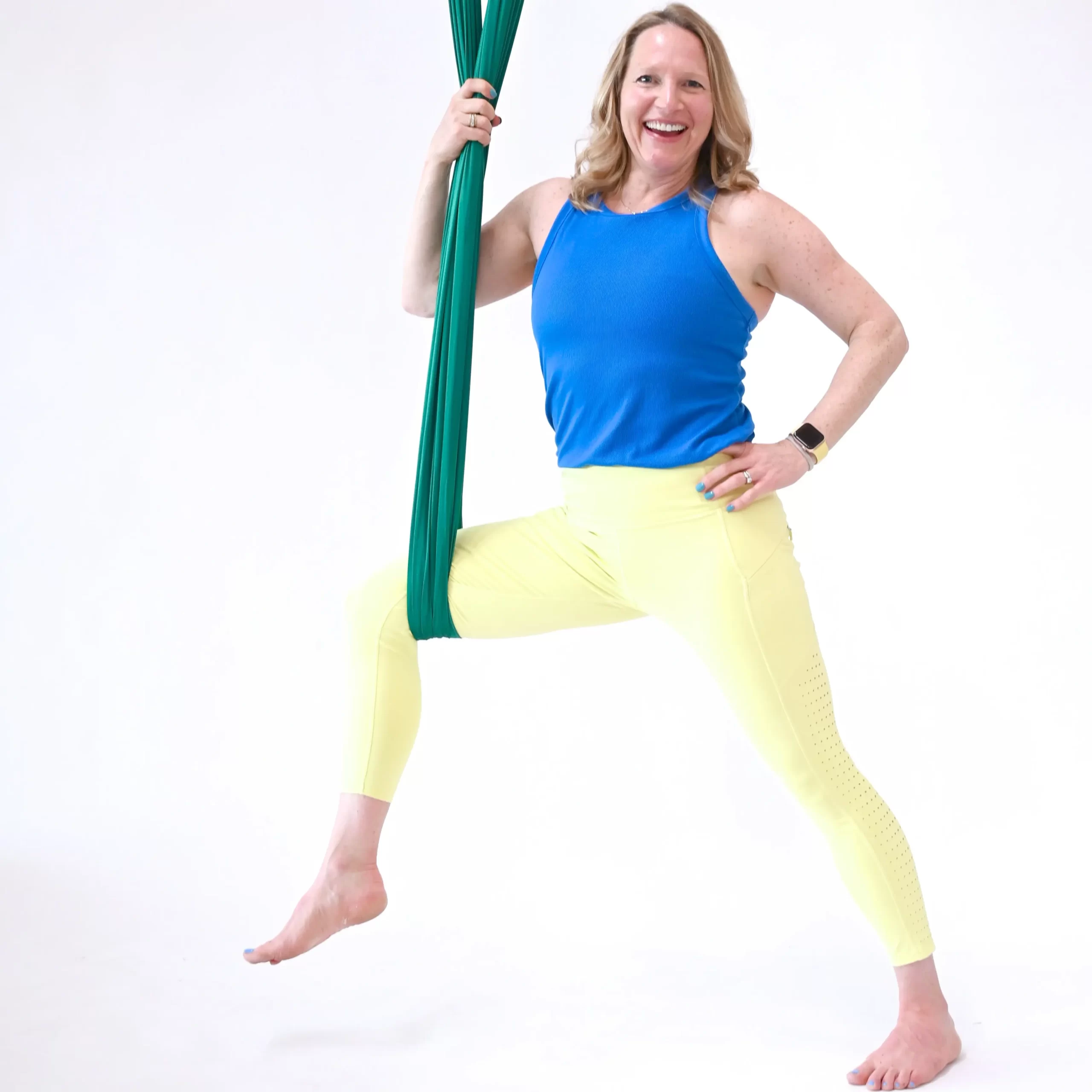 Yoga teacher is standing next to an aerial silk hammock with her right leg bent and being supported under her knee with the hammock. She is smiling and her left hand is on her left hip.