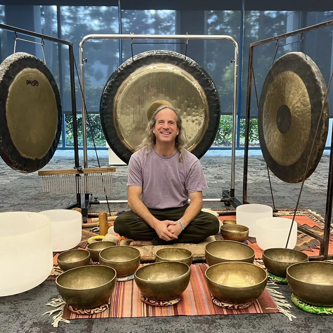 Danny Goldberg is seated with his legs crossed and his hands together near his center. Behind him are three brass gongs. On the ground in front of him, in a semi circle are crystals sound bowls and brass sound bowls.