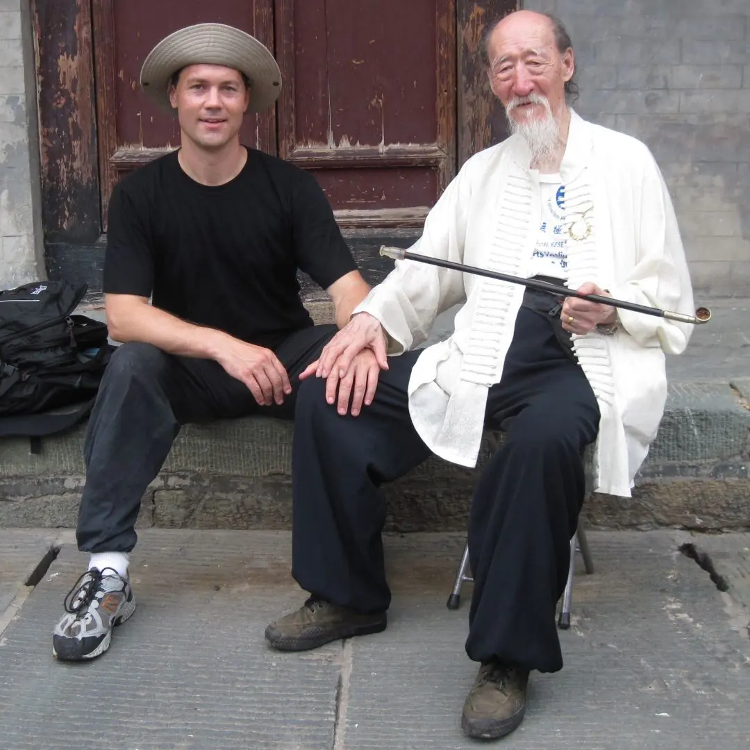 Two men sitting outdoors. The man on the left has his left hand on the other man's right knee. The man sitting on the right has his right hand on top of the other man's hand on his knee. The man on the right is holding a smoking pipe, approximately 2 feet long.