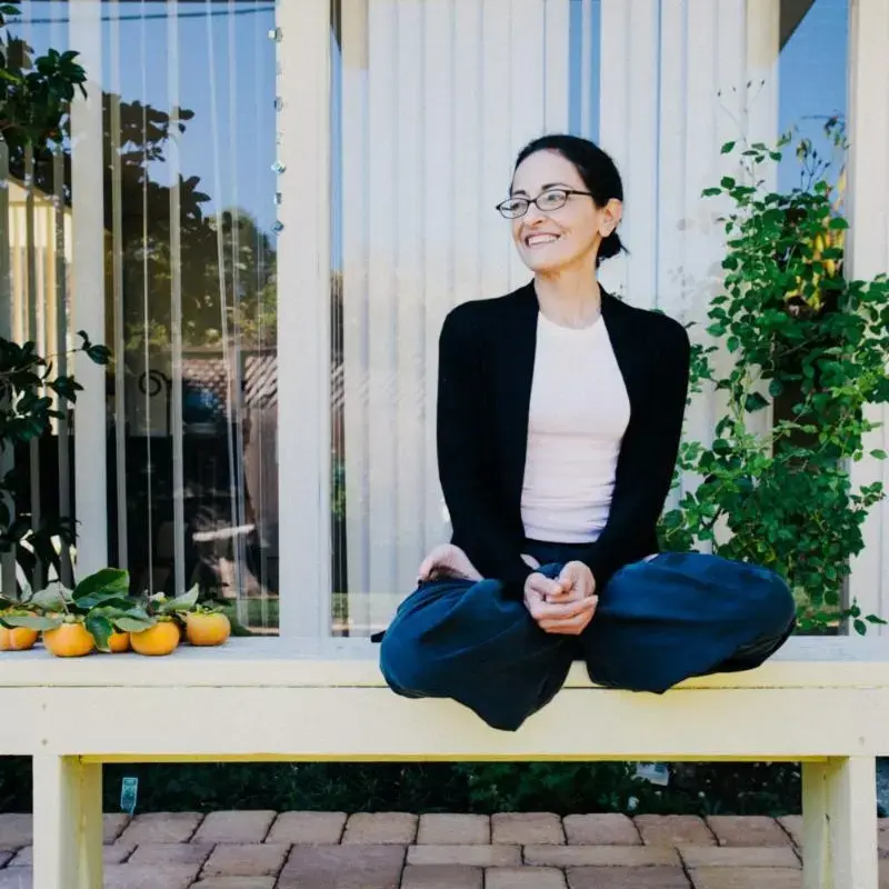 Mojdeh is sitting cross-legged on an outdoor bench, while smiling.