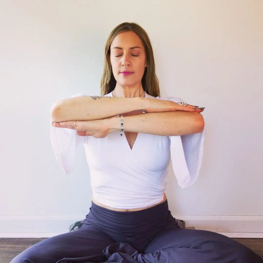 Naomi Wilder is seated cross legged on a floor. Her eyes are closed, while her arms are parallel with her forearms stacked at shoulder height.