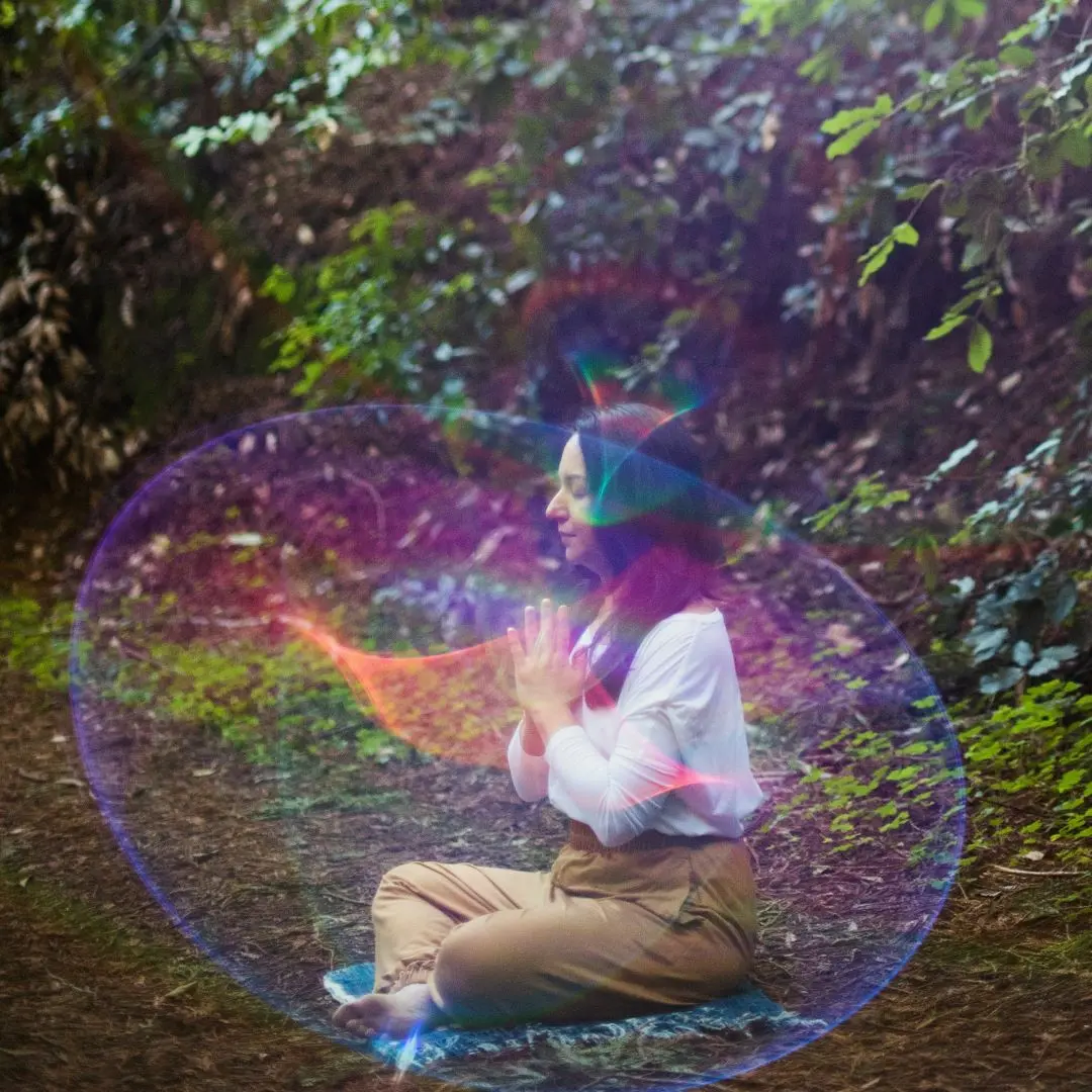 Paulette Sato sitting crossed legged on a folded blanket. Her arms are folded with her hands together in front of her heart. She is outside on dirt path, with green shrubbery behind her. There is an iridescent and translucent bubble over her.