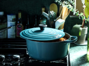 A turquoise crockpot sits on a stove with the lid open just a bit revealing food cooking inside. Light comes in from a window to the right and eucalyptus branches peak into frame from the right as well.