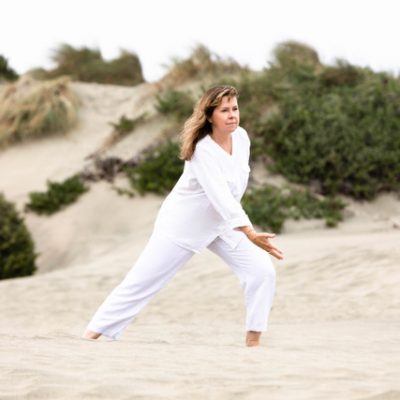 A yoga teacher dressed in all white, is practicing qigong on a beach with a sand dune and green shrubbery behind her.
