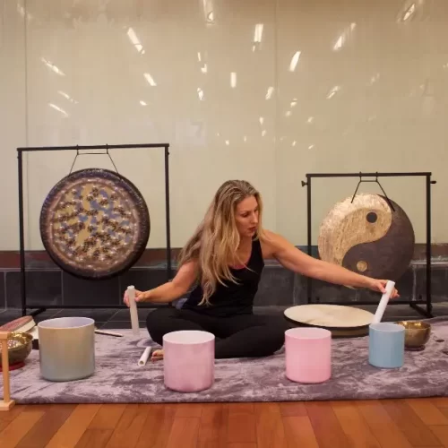 Erica Valentine is seated on the floor with playing two crystal sound bowls at the same time. Behind her a two gongs.