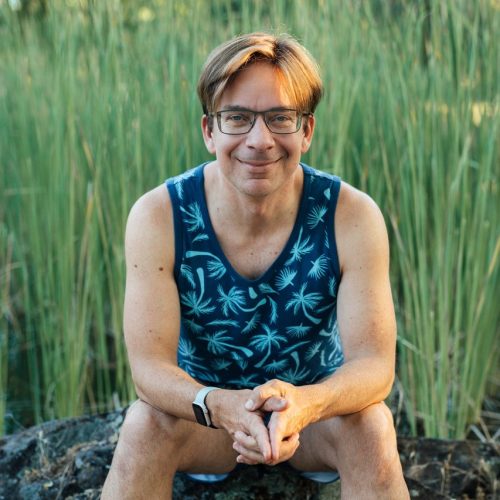 Jayson is sitting on a mossy rock with tall green reeds behind him.