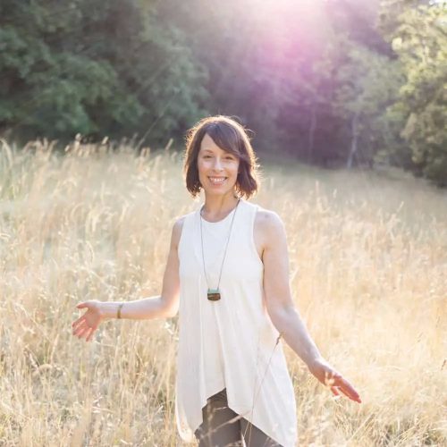 Jill Lacher standing in a field with tall, wheat colored plants. Her hands are at her hip height and lightly touching the plants. The sun is shining on her from behind.