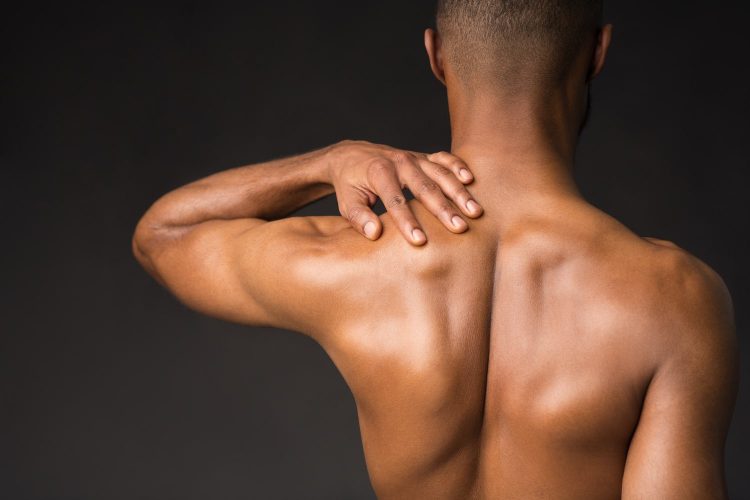 Rear view of afro man rubbing neck, suffering from arthritis, cervical chondrosis, massaging shoulder, black wall