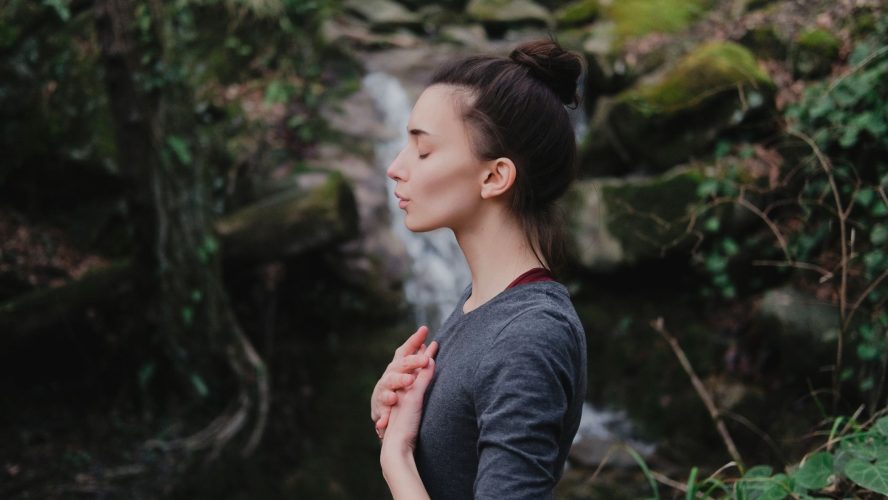 Young woman practicing breathing yoga pranayama outdoors in moss forest on background of waterfall. Unity with nature concept