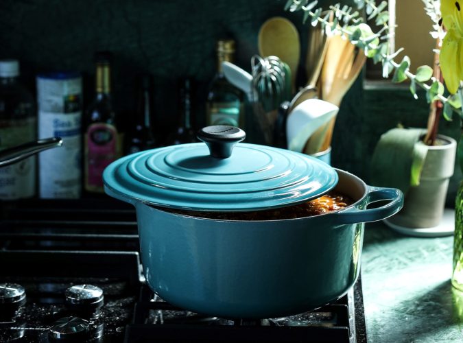 A turquoise crockpot sits on a stove with the lid open just a bit revealing food cooking inside. Light comes in from a window to the right and eucalyptus branches peak into frame from the right as well.