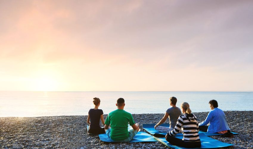 Group of people practicing morning meditation, yoga outdoors at sunrise on the beach, sitting and meditating in Padmasana exercise, Lotus yoga pose. Feeling so comfortable and relax. Healthy lifestyle.
