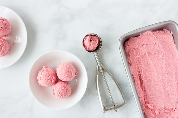 Homemade pink forced rhubarb ice cream in stainless steel tub, ice cream scoop and on plates. Top view.