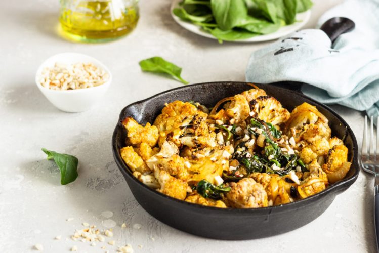 Roasted cauliflower with spinach, raisin and nuts in a cast-iron pan. Light grey background. Copy space. Healthy vegan food.