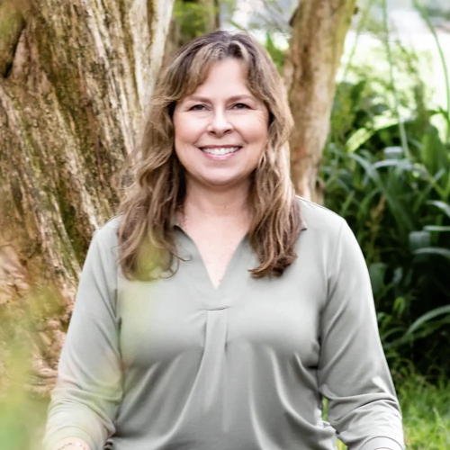 Yoga teacher, Joanne Varni, is smiling while seated outdoors, in front of a tree and green foliage.