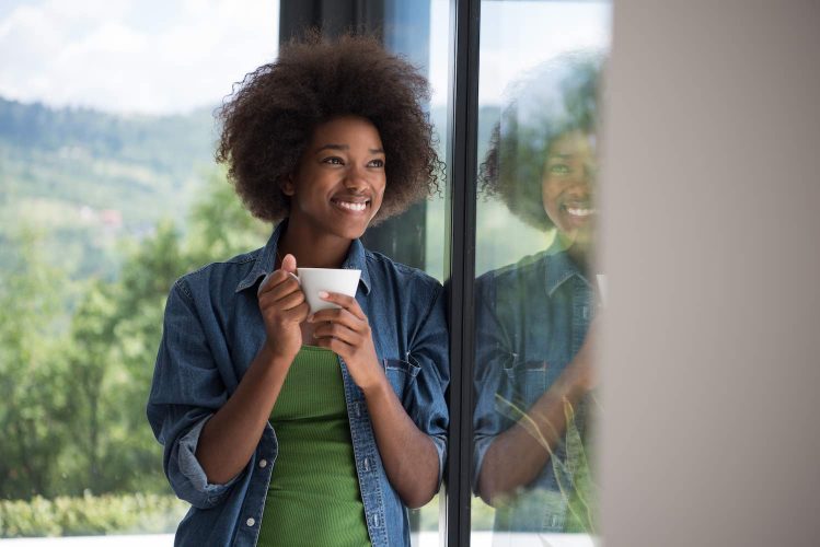 Beautiful young african american woman drinking coffee and looking through a window in her luxury home