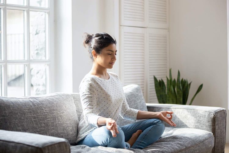 Indian female sitting on couch lotus pose put hands on lap folded fingers closed eyes enjoy meditation do yoga exercise at home. No stress, healthy habit, mindfulness lifestyle, anxiety relief concept