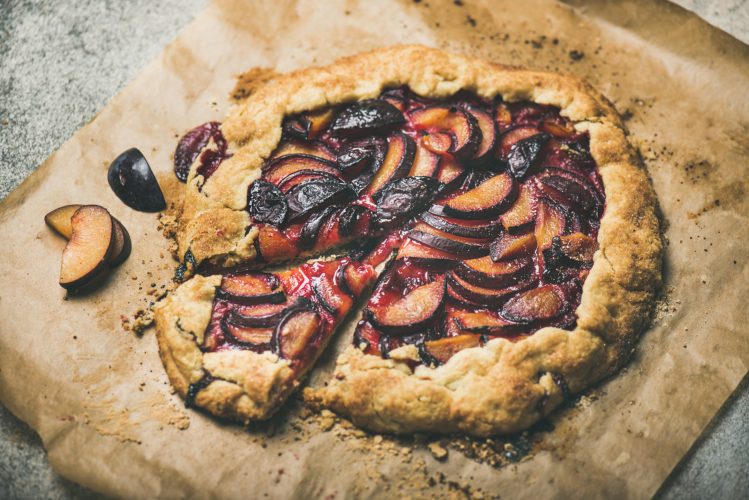 A plum galette sits on a wooden serving board. A slice is cut out at the bottom left. We see the dessert from above as if looking down at it.
