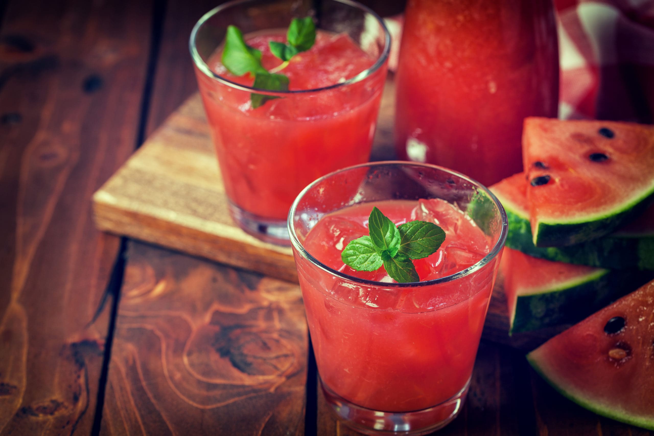 Two glasses filled with blended watermelon cooler and garnished with mint sit on a wooden table with watermelon slices in the background to the right.